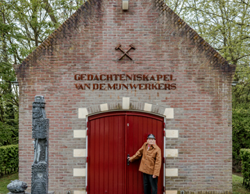 Oud-mijnwerker en oprichter van de gedachteniskapel poseert voor de rode deur van de gedachteniskapel.